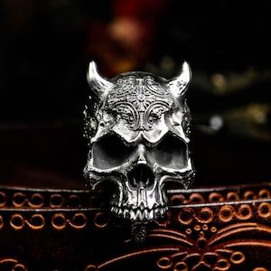 May include: A silver skull ring with intricate detailing, including horns and ornate patterns on the forehead. The ring has a gothic or biker aesthetic, with a dark, shadowy background.