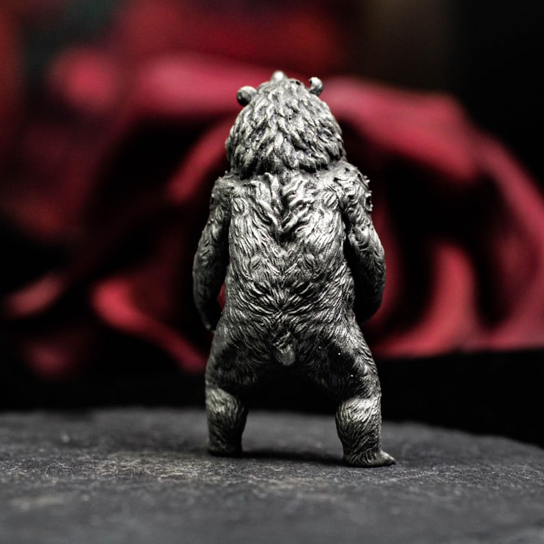 May include: A detailed, silver-colored bear figurine, viewed from the back. The bear has textured fur and stands on all fours. The background is a blurred dark red.
