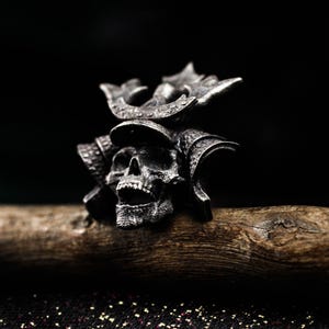 May include: A detailed silver skull ring with a samurai helmet design. The ring features intricate carvings and sits on a wooden surface against a dark background. The skull's mouth is open, and the helmet has decorative elements.