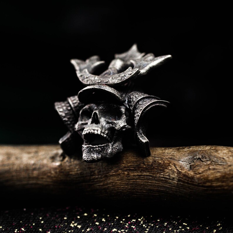 May include: A detailed silver skull ring with a samurai helmet design. The ring features intricate carvings and sits on a wooden surface against a dark background. The skull's mouth is open, and the helmet has decorative elements.