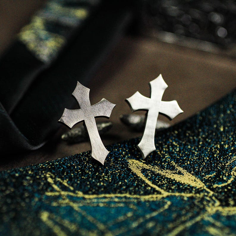 May include: A pair of silver-colored cross-shaped earrings. The earrings have a gothic design with pointed edges. They are displayed on a textured, dark blue and gold patterned surface.