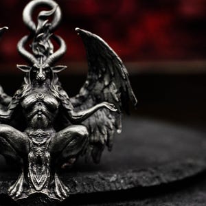 May include: A detailed, silver-toned sculpture of Baphomet, a goat-headed deity, with large wings and a seated posture. The sculpture has intricate details, including horns, a beard, and a flame above the head. The background is a dark red.