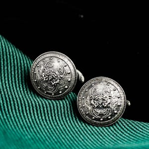 Puede incluir: Un par de gemelos plateados con un diseño floral y escudo detallado. Los gemelos redondos se muestran sobre una tela verde texturizada sobre un fondo negro. El intrincado diseño presenta un motivo floral central rodeado por un borde decorativo.