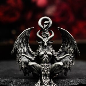 May include: A detailed, silver-colored pendant depicting Baphomet, a goat-headed deity, with large feathered wings and a flame above its head. The pendant is intricately designed, showcasing the figure's horns, facial features, and seated pose. The background is a blurred red.