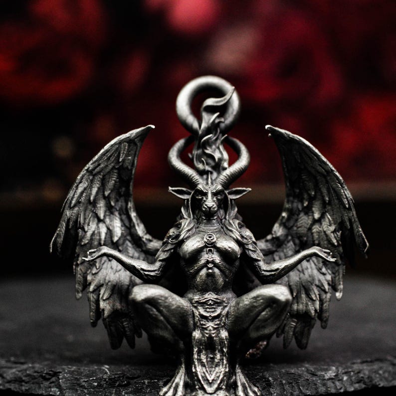 May include: A detailed, silver-colored pendant depicting Baphomet, a goat-headed deity, with large feathered wings and a flame above its head. The pendant is intricately designed, showcasing the figure's horns, facial features, and seated pose. The background is a blurred red.