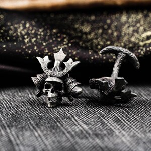 May include: A pair of silver-toned cufflinks. One cufflink features a skull wearing a samurai helmet, and the other has a textured design. The cufflinks are set against a patterned gray background.
