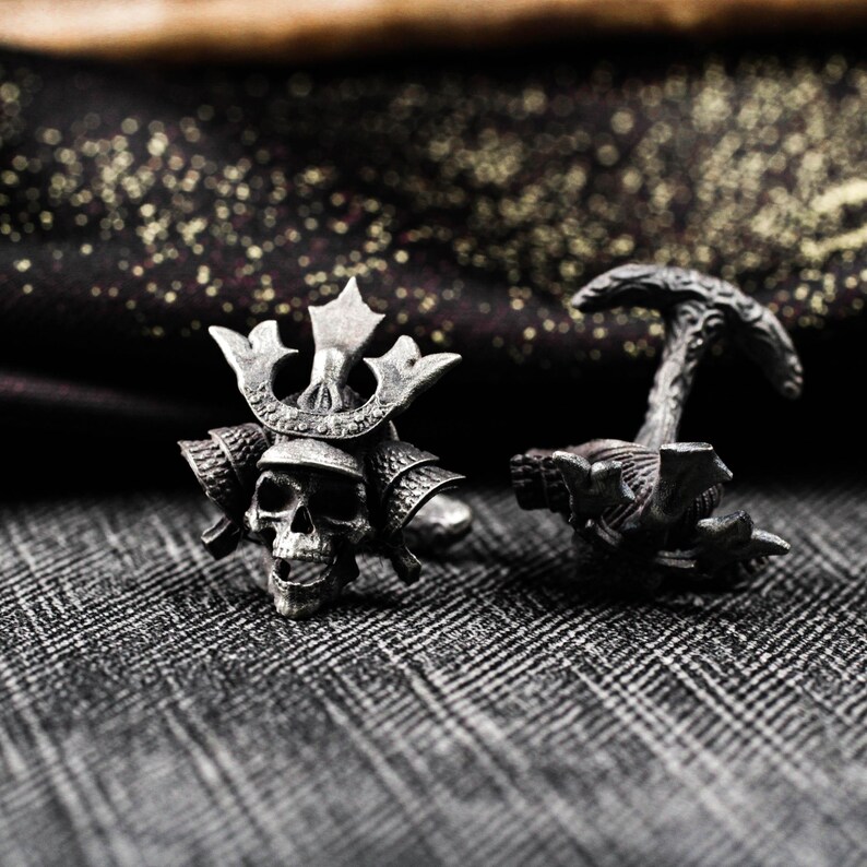 May include: A pair of silver-toned cufflinks. One cufflink features a skull wearing a samurai helmet, and the other has a textured design. The cufflinks are set against a patterned gray background.