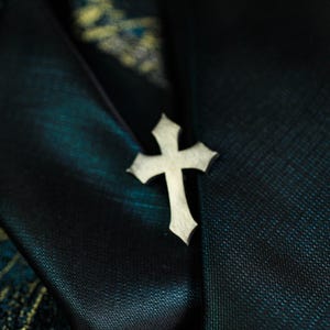May include: A silver-colored cross pin with a textured surface. The cross is set against a dark teal fabric with subtle patterns, creating a contrast. The pin is a religious symbol.