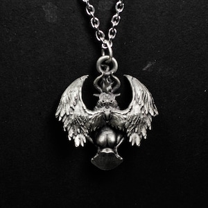 May include: Silver-toned pendant necklace featuring a detailed winged creature with horns and a flame above its head. The pendant is suspended from a delicate chain, creating a gothic or fantasy aesthetic. The design is intricate and three-dimensional.