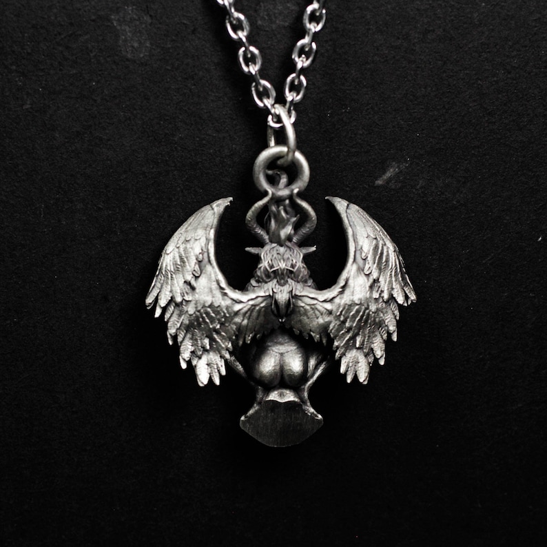 May include: Silver-toned pendant necklace featuring a detailed winged creature with horns and a flame above its head. The pendant is suspended from a delicate chain, creating a gothic or fantasy aesthetic. The design is intricate and three-dimensional.