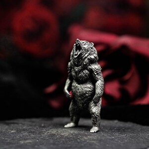 May include: A detailed silver-colored bear figurine in a standing pose with its mouth open, as if roaring. The bear has textured fur and is set against a blurred background of dark red and black.
