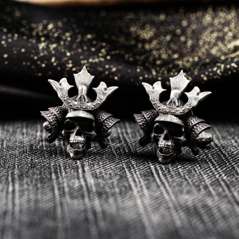 May include: Pair of silver-toned cufflinks featuring a skull design with a samurai helmet. The cufflinks have intricate details and a dark, oxidized finish. The background is a textured gray fabric.