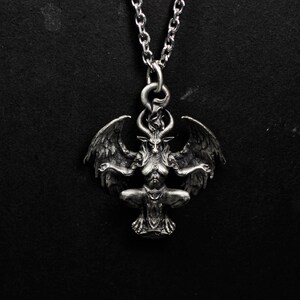 May include: A silver-colored pendant necklace featuring a detailed depiction of Baphomet. The pendant shows a winged, goat-headed figure in a seated pose, with intricate detailing on the horns, wings, and body. The necklace has a chain.