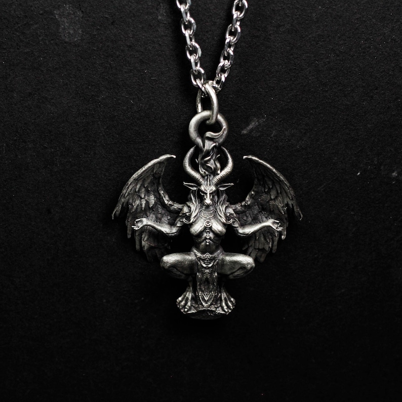 May include: A silver-colored pendant necklace featuring a detailed depiction of Baphomet. The pendant shows a winged, goat-headed figure in a seated pose, with intricate detailing on the horns, wings, and body. The necklace has a chain.