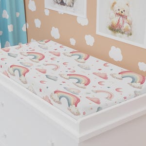 Könnte beinhalten: Ein weißer Wickeltisch mit einer Wickelauflage mit Regenbogenmuster. Die Auflage zeigt Aquarell-Regenbögen, Wolken und Sterne in Pastellfarben. Der Wickeltisch befindet sich in einem Kinderzimmer mit Teddybär-Kunst.