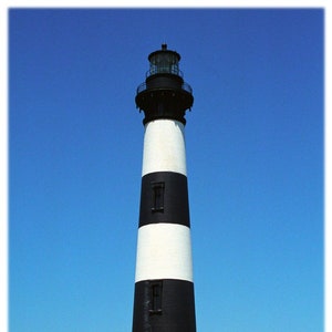 Op de afbeelding: Een zwart-wit gestreepte vuurtoren met een wit huis aan de basis. De vuurtoren is hoog en slank, met een zwart dak en een witte lantaarnkamer bovenop. Het huis is klein en rechthoekig, met een wit dak en een rode bakstenen schoorsteen. De vuurtoren staat tegen een helderblauwe lucht.