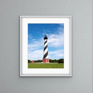 Op de afbeelding: Een ingelijste foto van een zwart-wit gestreepte vuurtoren met een rood dak tegen een blauwe lucht met witte wolken. De vuurtoren staat op een grasveld met bomen op de achtergrond.