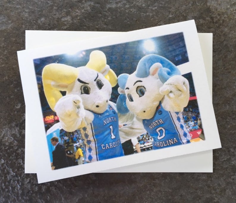 Puede incluir: Dos mascotas blancas que llevan camisetas azules y blancas con el texto "North Carolina" en ellas. Las mascotas son ambas carneros con cuernos azules y amarillos. La mascota de la izquierda est&aacute; apuntando con su mano derecha. La mascota de la derecha est&aacute; sonriendo y tiene su mano izquierda en el hombro de la mascota de la izquierda.