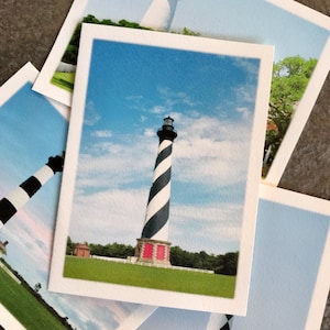 Op de afbeelding: Een zwart-wit gestreepte vuurtoren met een rood dak tegen een blauwe lucht met witte wolken. De vuurtoren is omgeven door groen gras.