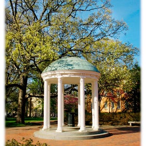 May include: A white, classical-style gazebo with a teal-colored dome roof stands in a park setting. The gazebo has several white columns and is surrounded by trees with green foliage and a brick-paved walkway. The sky is blue.
