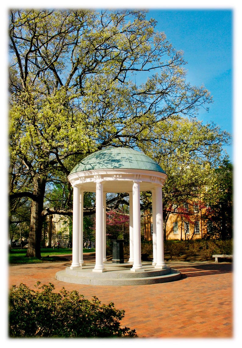 May include: A white, classical-style gazebo with a teal-colored dome roof stands in a park setting. The gazebo has several white columns and is surrounded by trees with green foliage and a brick-paved walkway. The sky is blue.