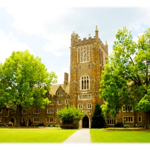 May include: A stone building with a clock tower and green trees in front of it. The building has a large arched entrance and many windows. The trees are in full bloom and the sky is blue.