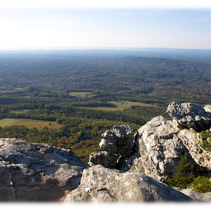 May include: A panoramic view from a rocky mountaintop, showcasing a vast expanse of forested hills and valleys. The foreground features large, gray, weathered rocks.