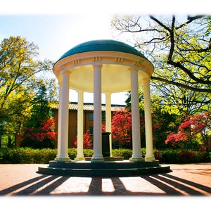 May include: A white, columned structure with a teal dome, set in a park-like setting. The building casts long shadows on the brick ground. Trees with green and red foliage surround the structure, under a bright blue sky.