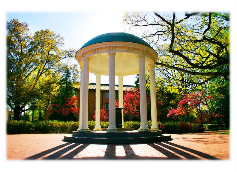 May include: A white, columned structure with a teal dome, set in a park-like setting. The building casts long shadows on the brick ground. Trees with green and red foliage surround the structure, under a bright blue sky.