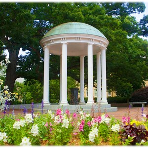 May include: A white, columned structure with a green dome roof stands in a garden filled with colorful flowers. The building is surrounded by lush green trees and a brick walkway. A bench is visible in the background.