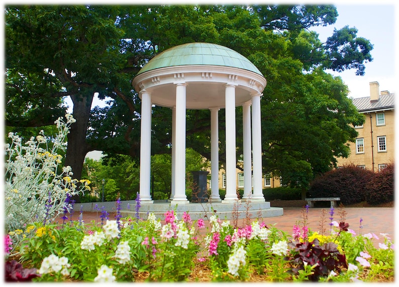 May include: A white, columned structure with a green dome roof stands in a garden filled with colorful flowers. The building is surrounded by lush green trees and a brick walkway. A bench is visible in the background.