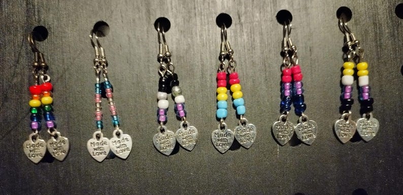 May include: A collection of beaded earrings with silver-toned heart charms that read "Made with Love." The earrings feature various color combinations, including rainbow, pink, blue, and black beads.
