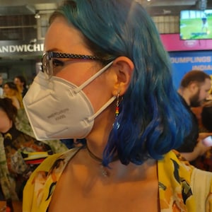 May include: A person wearing a white KN95 face mask with the text "wellbefore" and blue hair. They are wearing glasses and a yellow floral print top. Dangling earrings with colorful beads and feathers are visible.