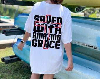 以下が含まれることがあります： 白地に黒と赤のグラフィックがプリントされたTシャツ。グラフィックには「SAVED WITH AMAZING GRACE」と書いてあり、十字架の形をしている。単語は積み重ねられている。