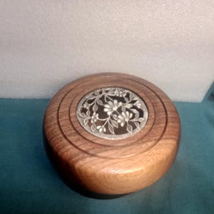 May include: A round wooden box with a decorative metal lid featuring a floral design. The box is made of dark wood with a natural finish.