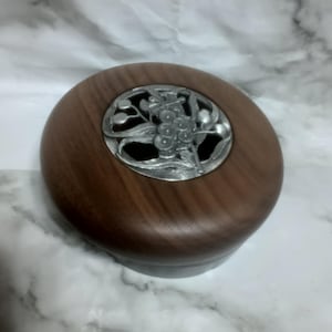 May include: A round wooden box with a dark brown finish and a smooth grain. The lid features a circular silver-toned metal inlay with an openwork design depicting a cluster of berries and leaves, centred on the top surface of the container.
