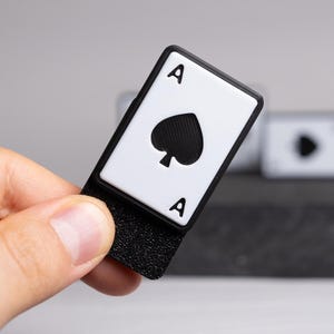 Può includere: Un segnalibro a forma di carta As di Picche in bianco e nero. Il segnalibro presenta il simbolo di picche e la lettera "A" in nero su sfondo bianco. La parte inferiore è nera e strutturata.