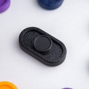 Pode incluir: Um spinner fidget preto com uma superfície texturizada. O spinner tem um botão central redondo preto.
