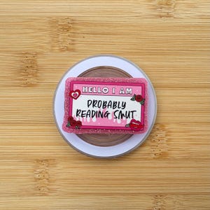May include: A pink and glittery badge with the text "HELLO I AM PROBABLY READING SMUT." The badge has a heart, roses, and a pair of lips. The badge is on a white and brown circular object.