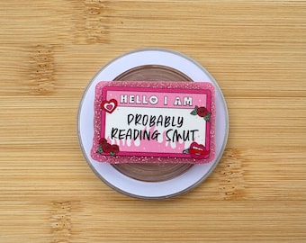 May include: A pink and glittery badge with the text "HELLO I AM PROBABLY READING SMUT." The badge has a heart, roses, and a pair of lips. The badge is on a white and brown circular object.