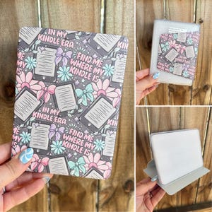 May include: A Kindle case with a gray background featuring a pattern of e-readers, flowers, and bows in pink, blue, and white. Text on the case reads "IN MY KINDLE ERA" and "FIND ME WHERE MY KINDLE IS".