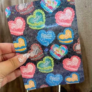 May include: A decorative sticker sheet featuring heart-shaped designs with various phrases. The hearts are in shades of blue, pink, green, and yellow, with phrases like "Happy Ever After" and "Enemies to Lovers". The background is dark with a speckled pattern.