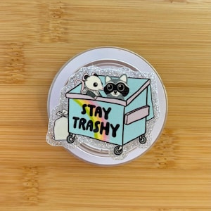 May include: A round, glittery phone grip featuring a light blue dumpster with a raccoon and a rat inside. The text "STAY TRASHY" is displayed on the front of the dumpster. A white trash bag is also visible.