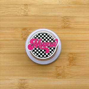 Può includere: Un supporto per telefono rotondo e bianco con la scritta "Mama" in rosa, con un carattere retrò. Il design include un motivo a scacchi in bianco e nero. Il supporto è su uno sfondo di legno marrone chiaro.