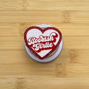 May include: A heart-shaped accessory featuring a red glitter border and a white background with a faint pink checkered pattern. The text Bookish Girlie is printed in a bold, red, retro-style font across the center of the heart-shaped surface.