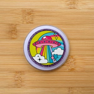 May include: A round, white pop-up phone grip with a colorful graphic of a pink UFO with heart-shaped windows, beaming a rainbow down to earth. The design includes clouds, stars, and a glittery purple border.