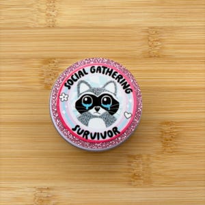 May include: A round, glitter-edged button with the text "SOCIAL GATHERING SURVIVOR" encircling a cartoon raccoon with teary eyes. The design features a pink and blue color scheme with a flower and heart accent.