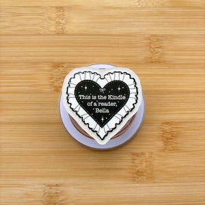 May include: A heart-shaped pop socket with a black center and white lace trim. The text "This is the Kindle of a reader, Bella" is printed on the front. The pop socket is on a light purple base.