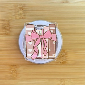 Op de afbeelding: Een popsocket met een stapel boeken vastgebonden met een roze strik. De boeken hebben een lichtroze kaft met witte hartjes, een witte kaft en een bruine kaft met witte bloemen. De popsocket staat op een witte basis.