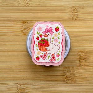 Op de afbeelding: Een decoratieve sticker met een cartoon gans die een mand met aardbeien draagt. De gans draagt roze laarzen en een roze strik. De sticker heeft een roze rand met aardbeien- en bloemaccenten. De achtergrond is een lichte houtnerf.